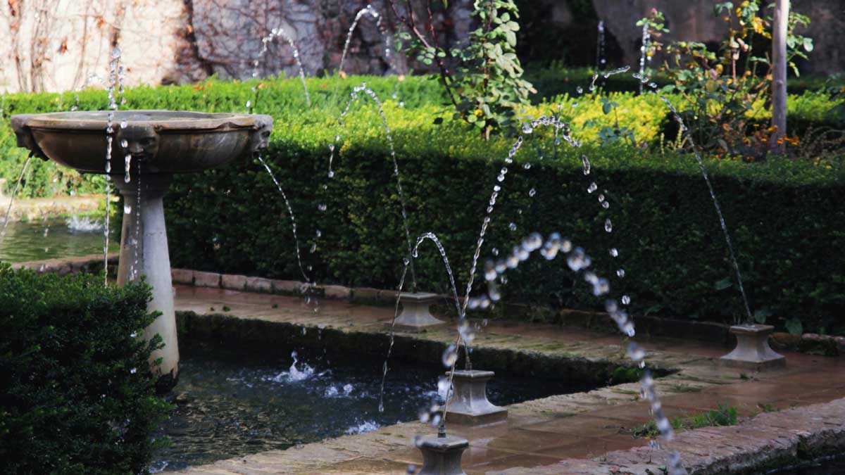 Classical stone fountain with multiple water jets in a serene garden setting, surrounded by lush hedges and plants.
