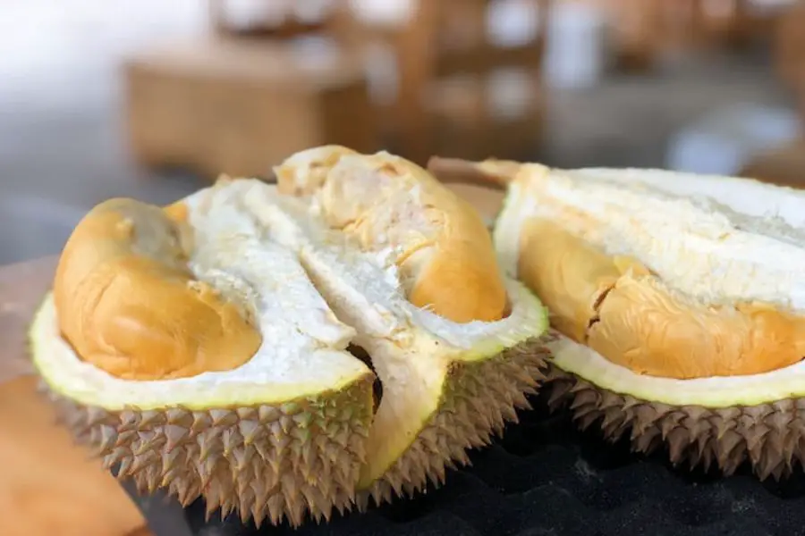 Open durian fruit revealing its soft yellow flesh, with spiky green shell, on a wooden surface.