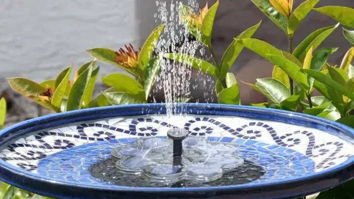 Water gently cascades from a solar-powered fountain in a blue and white mosaic-tiled bird bath, flanked by green plants with red tips.