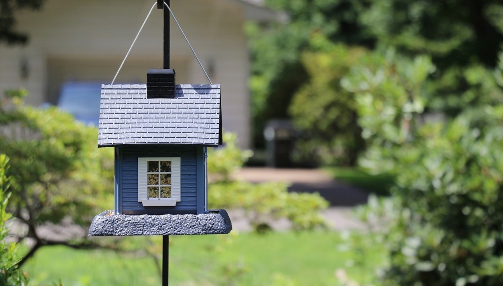 Quaint blue birdhouse with shingled roof hangs in garden setting.